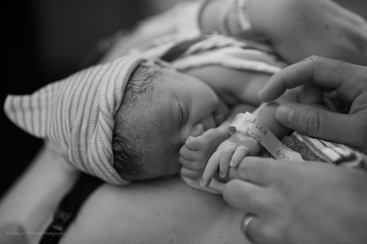Newborn resting skin-to-skin while an adult gently holds the baby's hand